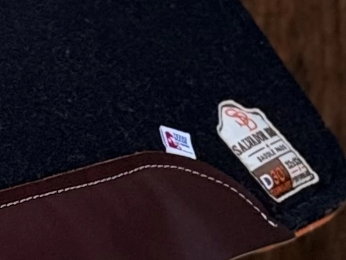 The rear corner of a Salvador Bros pad: black wool felt, the orange edge binding, the leather wear strip and the embroidered Salvador Bros shield.